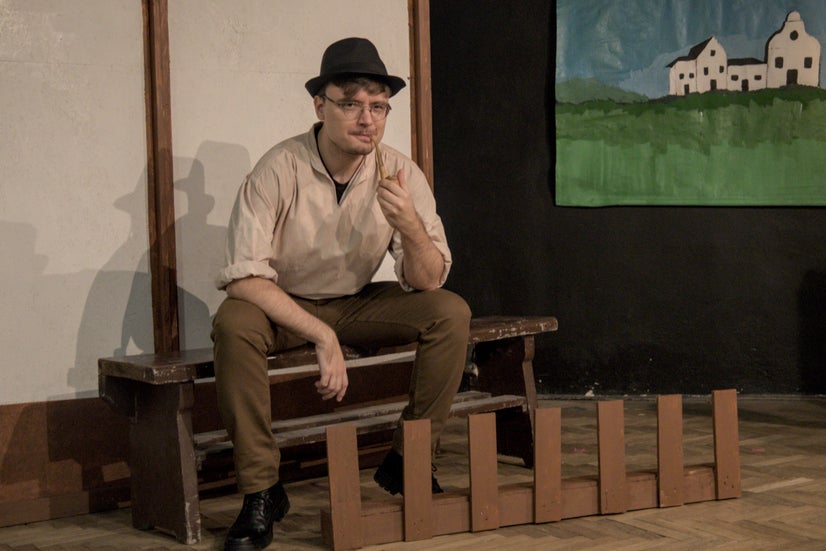 A man in an old-fashioned white shirt with a black hat sitting on a bench behind a small fence smoking a pipe.