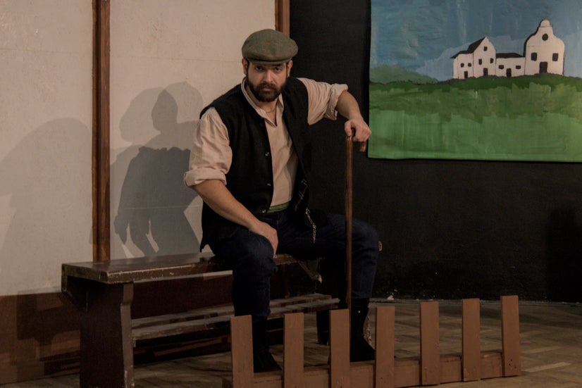 A man in an old-fashioned white shirt with a dark vest and a grey cap staring menacingly at the camera while sitting on a bench and resting on a cane.