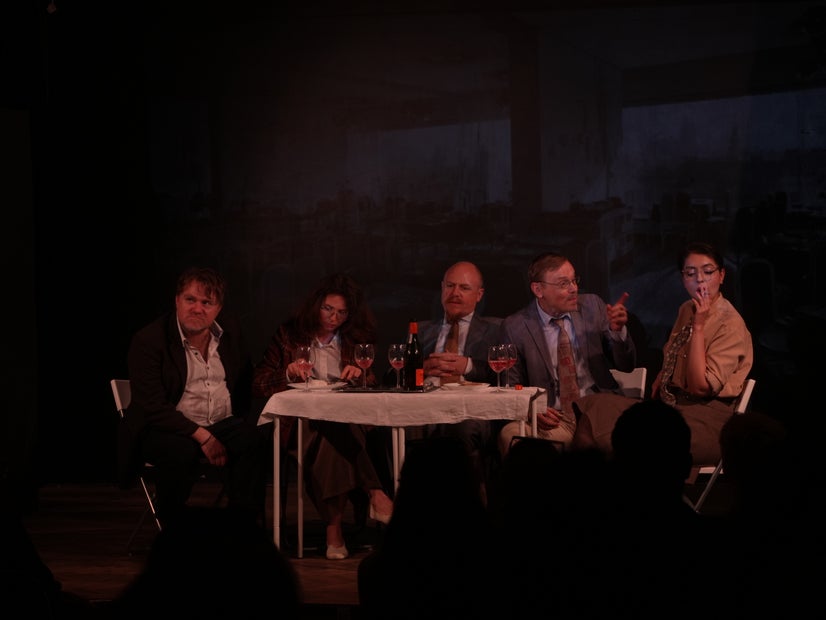 A table with food and drinks. Around it, a man looks annoyed, a woman is eating, a man looks sad, and another man is gesturing emphatically at a woman who is smoking.