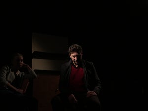 A man sits in the middle of a dark space with a light on his face. A woman sits nearby in shadow.