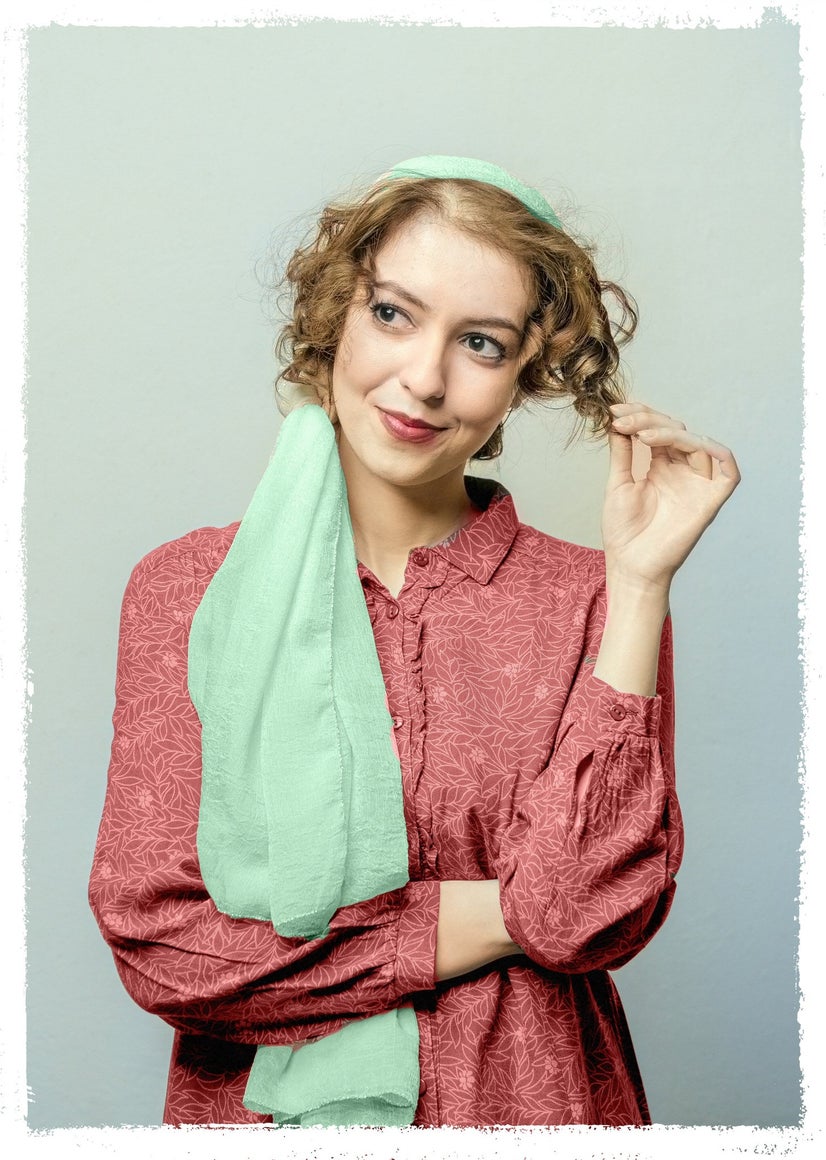 A woman in a red blouse with a bright teal scarf around her head and over her shoulder looks flirtatiously away from the camera while twirling her hair.