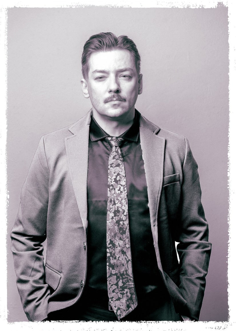 A man in a buttoned shirt, tie, and jacket stands with his hands in his pockets.