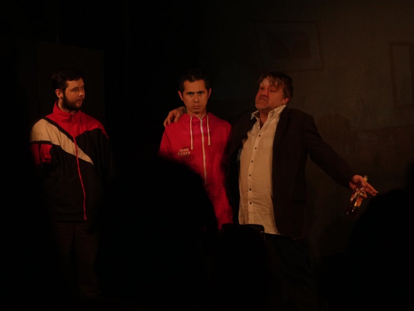 A man stares straight ahead in anger while another man with a cigarette and bottle in his hand hangs on the first man's shoulder. A third man looks on.
