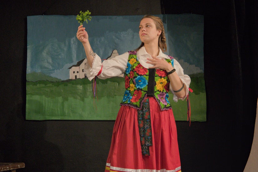 A woman in an old-fashioned white shirt with a top covered in flowers and a red skirt holding and looking at a sprig of parsley.
