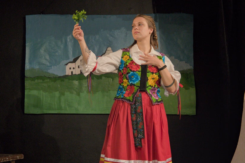 A woman in an old-fashioned white shirt with a top covered in flowers and a red skirt holding and looking at a sprig of parsley.