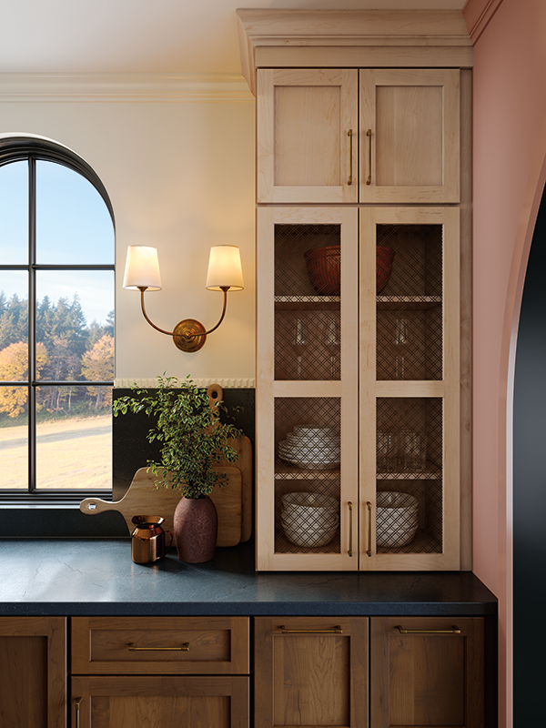 The mood of this image is warm, aglow from the kitchen sconce to the left of the cabinets. On the right, kitchen cabinets are closed, but wire-front mesh details are in the place of wooden panel doors, displaying peeks of the bowls and glassware from within.