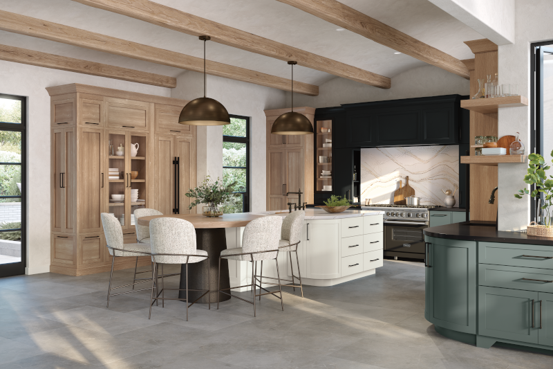 A round wooden table, with softly textured seating, creates a welcoming open kitchen design, complete with white kitchen island, nearby wooden hutch, and dark accent wall at the back painted black. This space is filled with natural lighting from the left, and has plants set out on each surface.