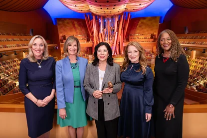 Five supervisors stand side by side in a brightly lit concert hall with colorful patterned seating and a large abstract wooden structure rising behind them. From left to right, Supervisor Kathryn Barger, Supervisor Janice Hahn, Supervisor Hilda Solis (Chair), Supervisor Lindsey Horvath, Supervisor Holly Mitchell. They wear formal attire including dresses, a blazer over a green dress, a checked blazer with a gavel in hand, a navy satin dress, and a black dress with a red necklace that reads 'love'; warm lighting highlights the wooden architectural details and curved ceiling panels throughout the hall.
