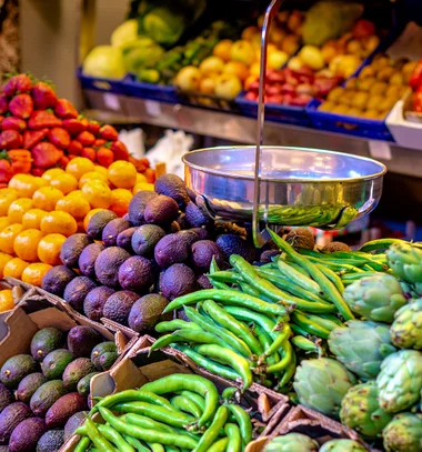 Colorful produce display at a market showing piles of avocados, green chili peppers, artichokes, tangerines, and strawberries. A metal hanging scale is positioned above the fruits and vegetables, with additional produce visible in the background.