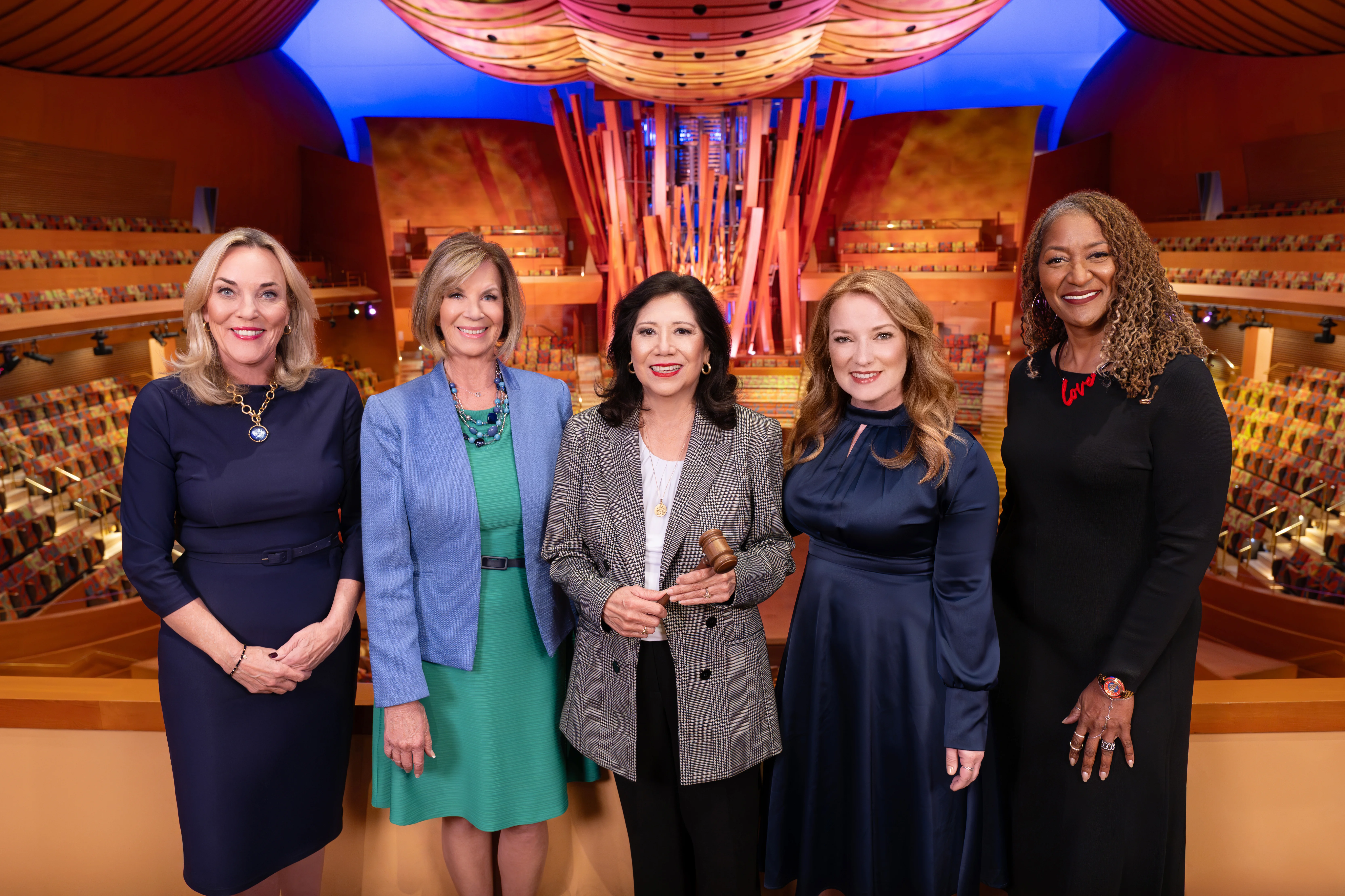 The 5 LA County Supervisors stand side by side inside a large auditorium or concert hall with rows of colorful seats and a large pipe organ structure in the background. The current chair (Hilda Solis), in the center holds a wooden gavel. The interior features warm lighting, curved architectural elements, and a decorative ceiling with circular light fixtures.