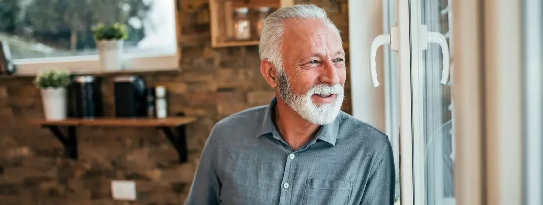 Man drinking coffee and looking through window, at home. Medigap Health Supplements.