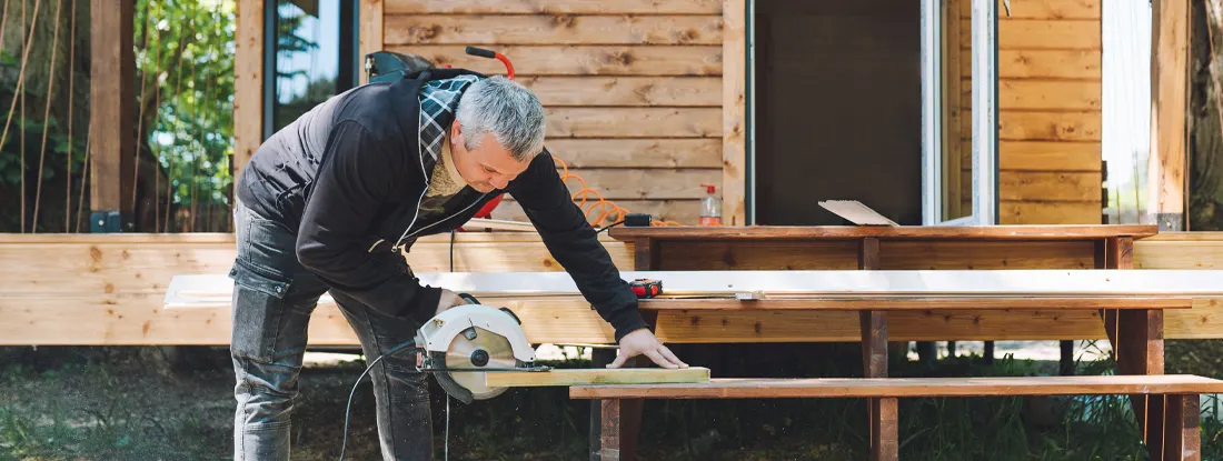 Carpenter operating circular saw grinder to cut wooden board for log cabin. Find Workers Comp for Self-Employed.