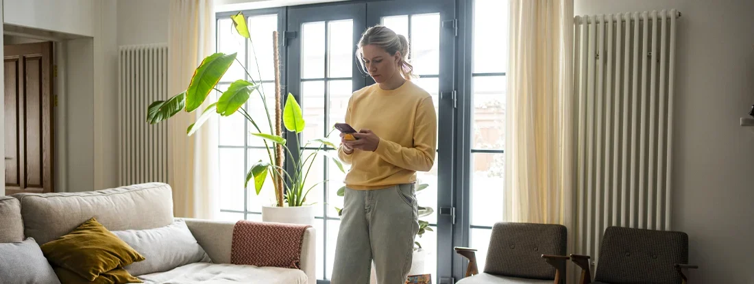 Woman in a yellow sweater using a smartphone in a cozy living room with plants. 10 Tips for Buying a House on Contract.