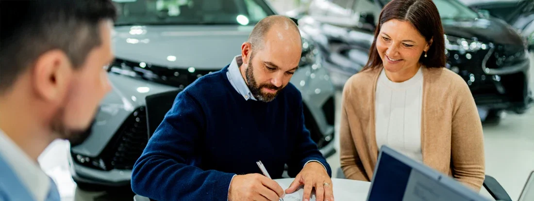 Couple signing documents at car dealership. Car Buyer Fees 101: Insurance, Taxes and Tabs.