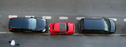 A small red car parked between two large black SUVs.
