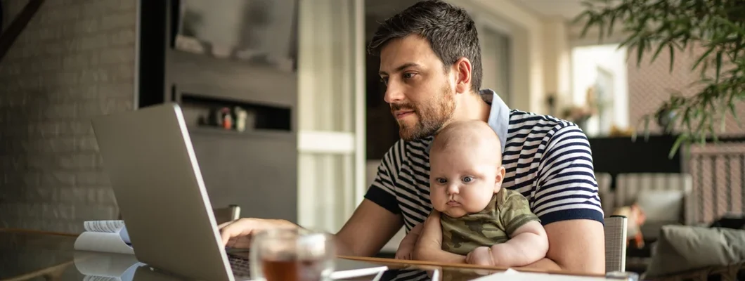 Man holding his newborn baby son and working with laptop at home. Find South Carolina Life Insurance.