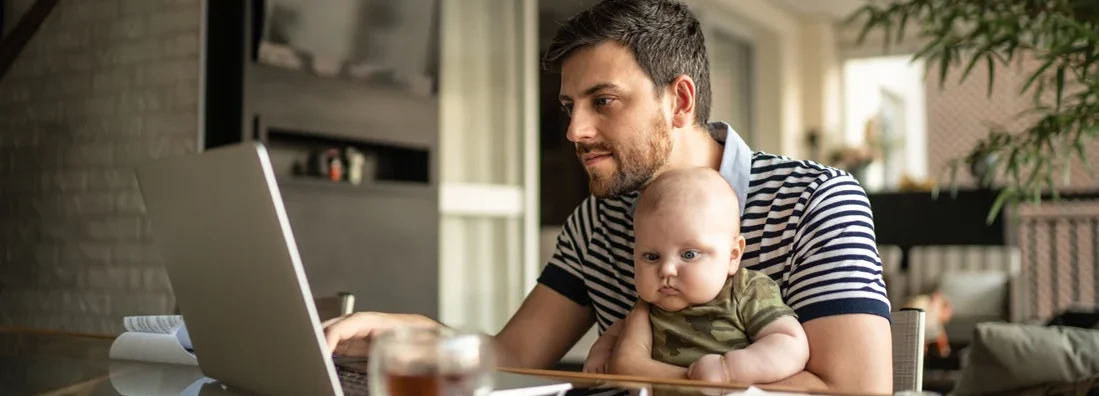 Man holding his newborn baby son and working with laptop at home. Find South Carolina Life Insurance.