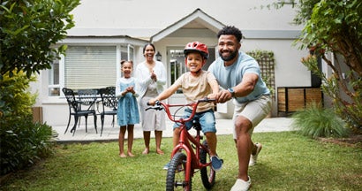 Proud dad teaching his young son to ride a bike while wearing a helmet in the backyard of their home. HO3 vs HO6 Insurance. 