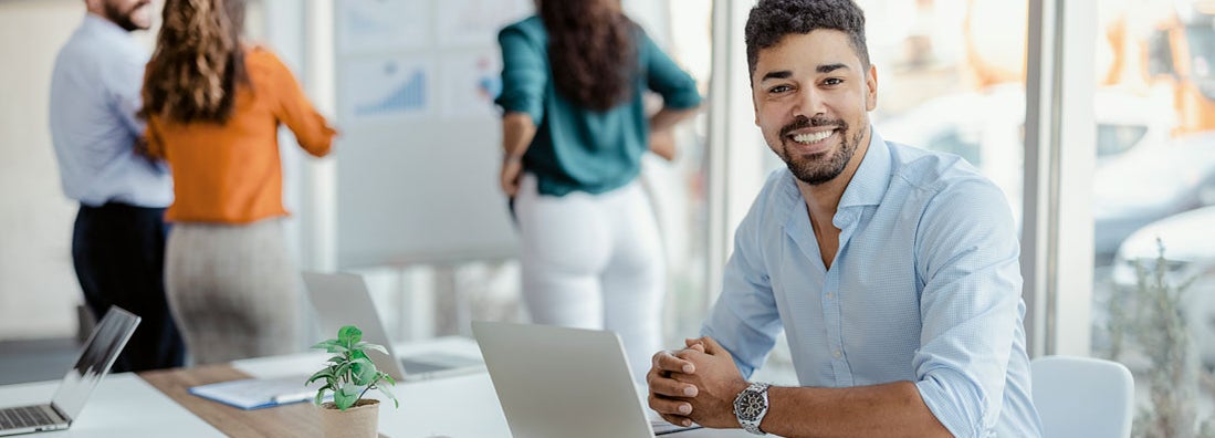 Smiling working man looking at camera with colleagues in the background. Find Business Umbrella Insurance. 