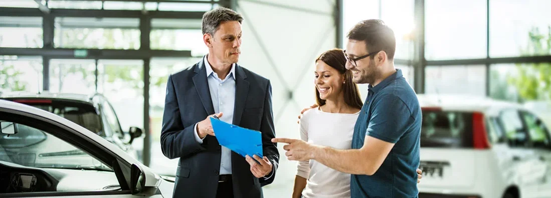 Couple having a conversation with a salesperson at a car dealership, all of them standing next to new cars. Find Car Dealer Insurance.