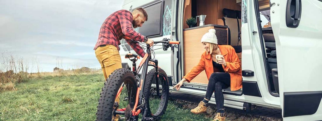 Woman drinking coffee sitting at the door of an RV and man checking bike. Find Massachusetts RV insurance.