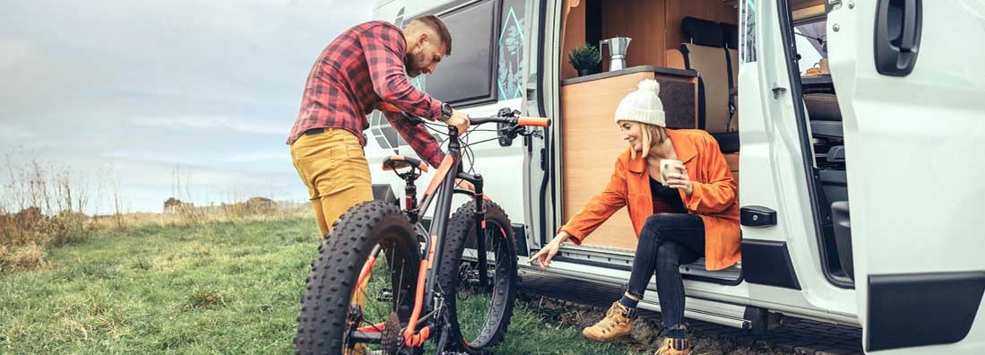 Woman drinking coffee sitting at the door of an RV and man checking bike. Find Massachusetts RV insurance.