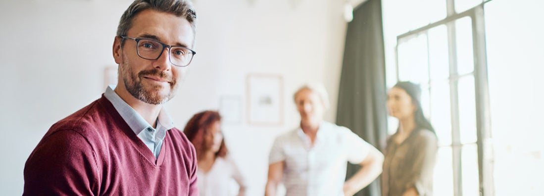 Businessman looking at the camera while working on his tablet and sitting in the office with employees in background. Find group disability insurance.