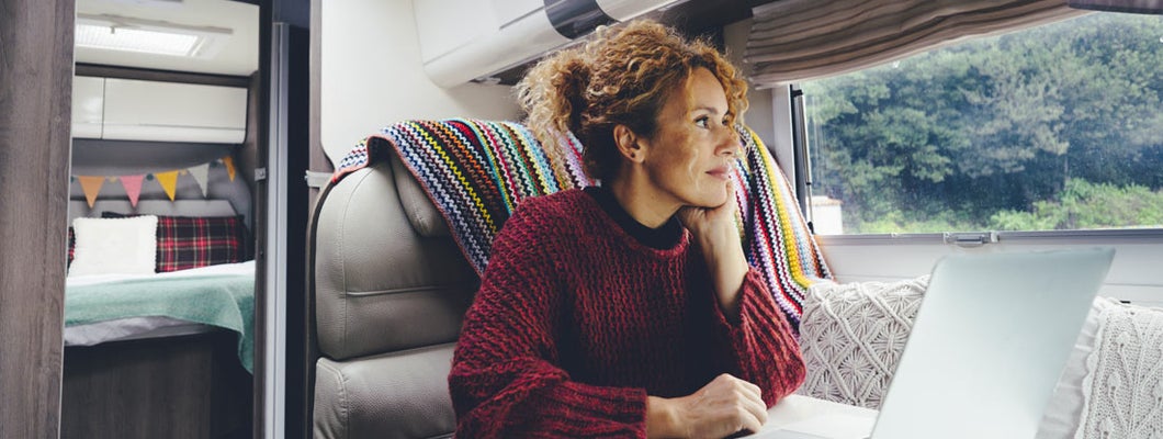 Woman using laptop inside a recreational vehicle sitting at the table with bedroom in background and nature park outside the windows. Find Vermont RV insurance.