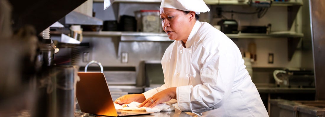 Chef and restaurant owner in commercial kitchen using laptop. Find Greensboro, North Carolina business insurance.