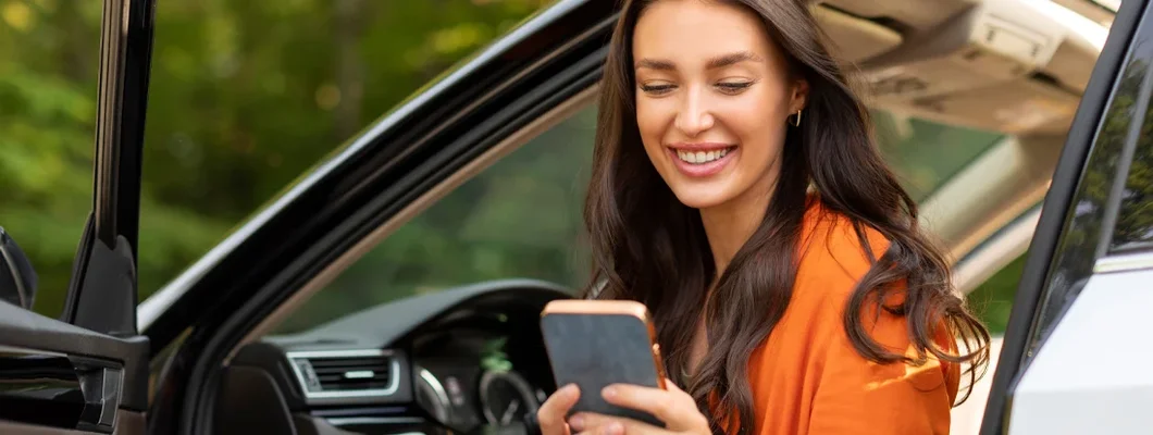 Woman using phone and sitting in car with open door. Why No Deposit Car Insurance May Be More Expensive.