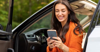 Woman using phone and sitting in car with open door. Why No Deposit Car Insurance May Be More Expensive.
