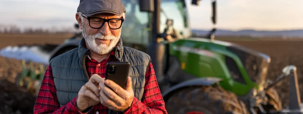 Senior farmer with mobile phone in field with tractor in background. Helpful Tips for Buying New or Used Farm Equipment. 
