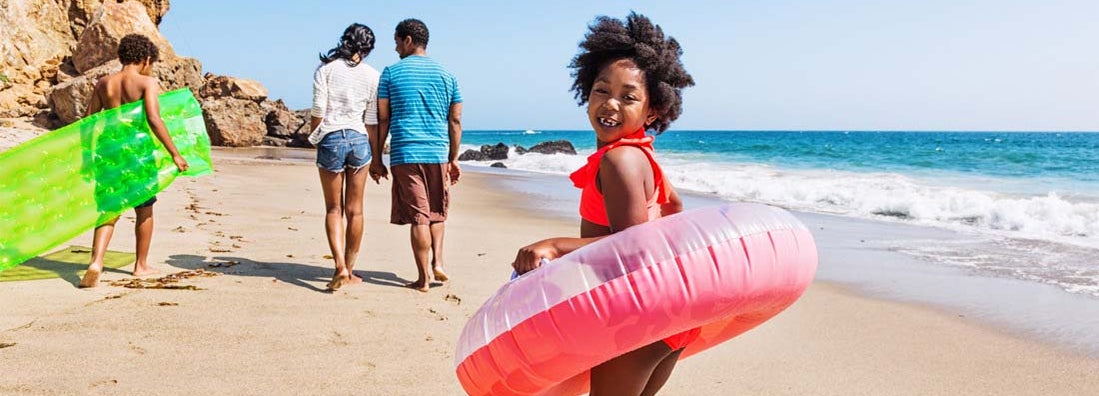 Family of four enjoying a day at the beach, walking with inflatable toys. Find term life insurance.