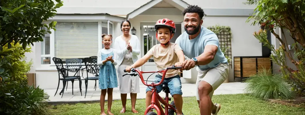 Proud dad teaching his young son to ride a bike while wearing a helmet in the backyard of their home. HO3 vs HO6 Insurance. 