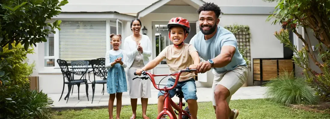 Proud dad teaching his young son to ride a bike while wearing a helmet in the backyard of their home. HO3 vs HO6 Insurance. 