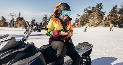 Woman sitting on snowmobile on hill of a snowcapped mountain using smart phone. Should I Get a GPS for My Snowmobile?