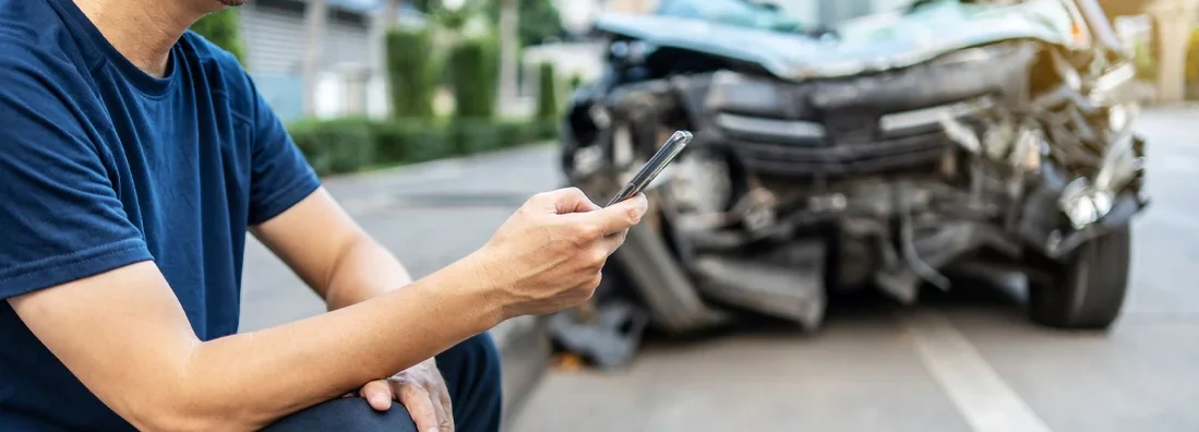 Man using phone to call his insurance agent after traffic accident with a background of the crashed car. How to file Car Insurance Claims.