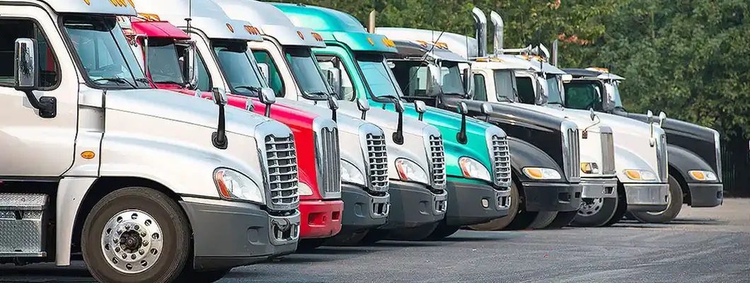 Different Big rig semi trucks tractors stand in row on parking lot with green trees. Find Owner Operator Insurance.