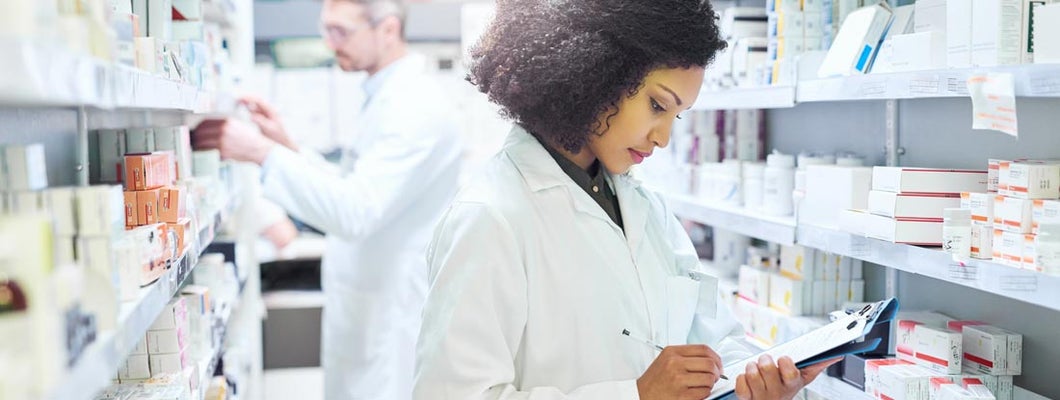 Pharmacist doing inventory in a pharmacy with her colleague in the background. Find Pharmacy Insurance.