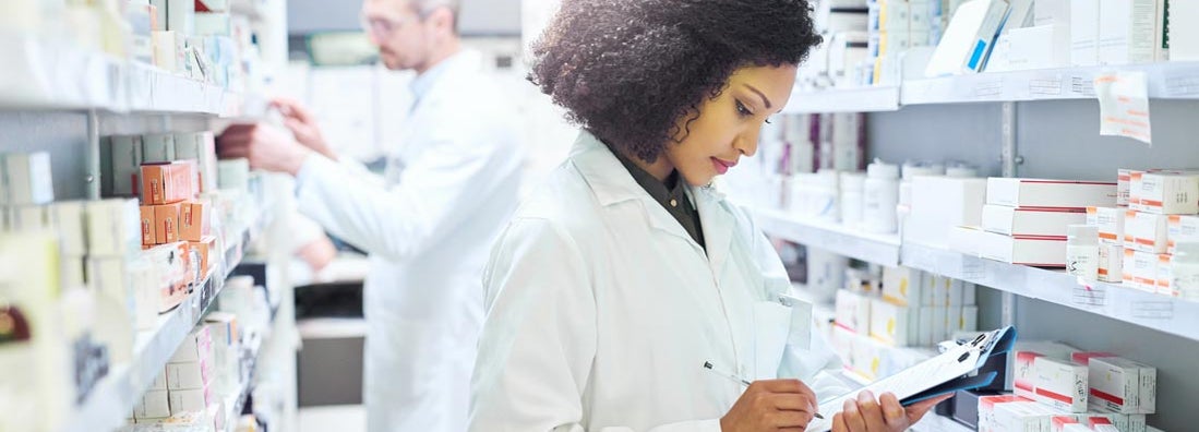 Pharmacist doing inventory in a pharmacy with her colleague in the background. Find Pharmacy Insurance.