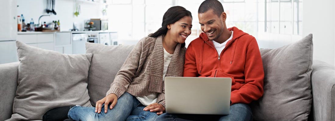 Smiling young couple sitting on their sofa using a laptop. Car Insurance Reviews.