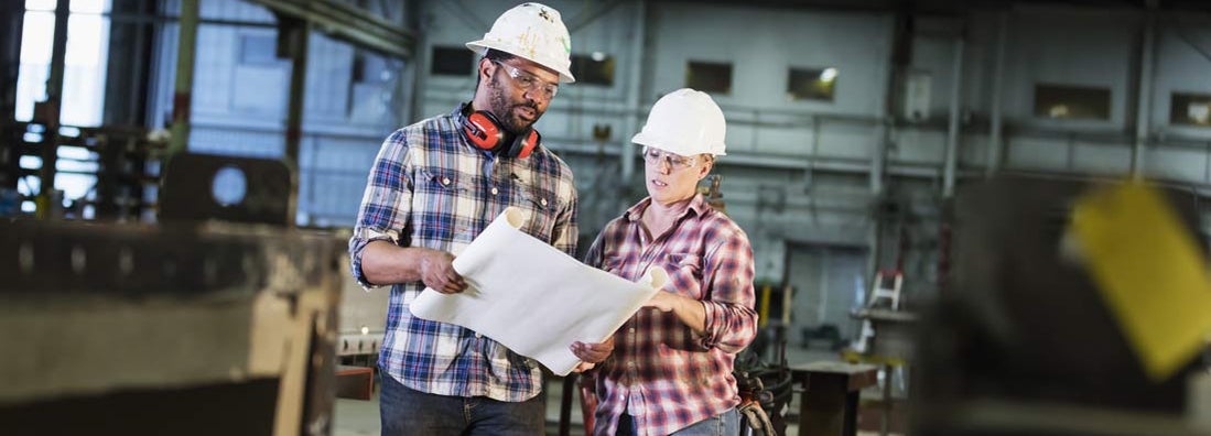 Man and woman standing on factory floor wearing hardhats and safety goggles, reading a set of plans. Self insured companies.