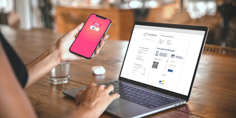 Une femme tenant un smartphone dans sa main se tient devant un ordinateur portable. Sur le smartphone, l'application Payconiq by Bancontact est ouverte, et sur l'ordinateur portable, un code QR Bancontact est visible pour finaliser le paiement.