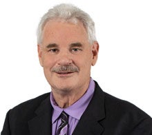 A man with gray hair, a mustache and wearing a purple shirt with a dark suit and tie.