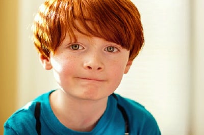 A young red-headed boy about 7 years old with hazel eyes looks directly at the camera with slight smile.