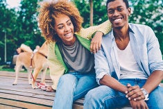 Young black couple, wearing blue jeans with their do on a leach are laughing while sitting on a wooden deck.