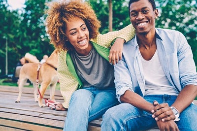 Young black couple, wearing blue jeans with their do on a leach are laughing while sitting on a wooden deck.