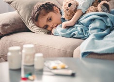 A young child lies under a blanket with a teddy bear looking at medications on the coffee table in front of her.