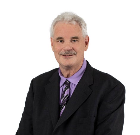 A close up a of man smiling at the camera with gray hair and a mustache wearing a purple shirt, dark suit and tie.