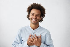 An African American man in his mid-thirties smiles at the camera while holding both hands over his heart.